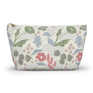 May include: A cream-colored zippered pouch with a floral pattern. The design features stylized flowers and leaves in shades of green, blue, pink, and gray, set against a backdrop of small gray dots. The zipper is gold-toned.