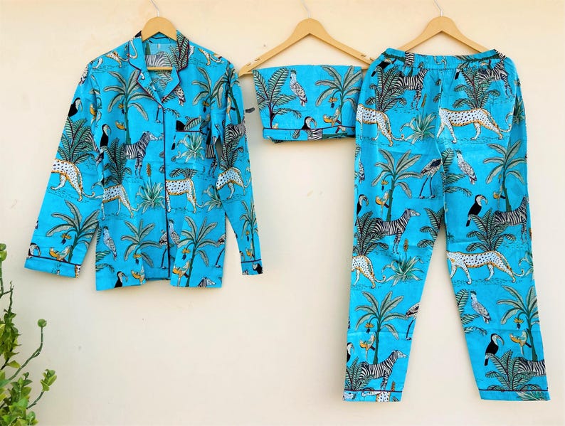 May include: A light blue pajama set with a jungle animal print. The set includes a button-down shirt and pants, both featuring zebras, leopards, toucans, and palm trees. The pajamas are displayed on wooden hangers.