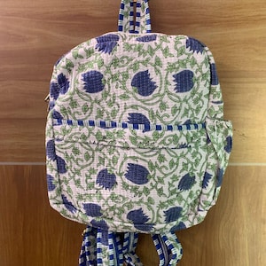 Puede incluir: Una pequeña mochila con un estampado floral. La mochila tiene un fondo blanco con diseños florales azules y verdes. Las correas y los ribetes son de rayas azules y blancas. La mochila está colgada de un gancho.