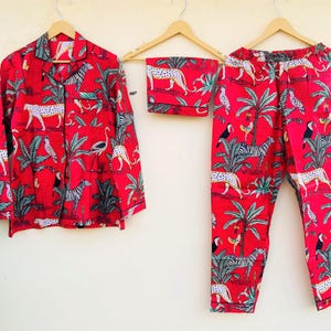 May include: Red pajama set with a jungle animal print, including zebras, leopards, and birds. The set includes a button-down shirt, shorts, and pants, all hanging on wooden hangers. The fabric features a vibrant red background.