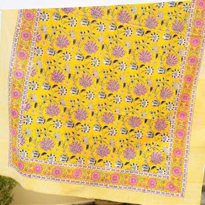 May include: A rectangular fabric textile featuring a bright yellow background with a repeating floral pattern in shades of pink and dark gray. The fabric has a wide border with a pink circular floral motif and thin yellow striped edges.