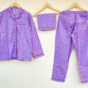 May include: A three-piece pajama set in a lavender hue, featuring a button-down shirt, shorts, and pants. The set is adorned with a repeating pattern of small figures in blue and pink. The edges are trimmed with a dark red piping.