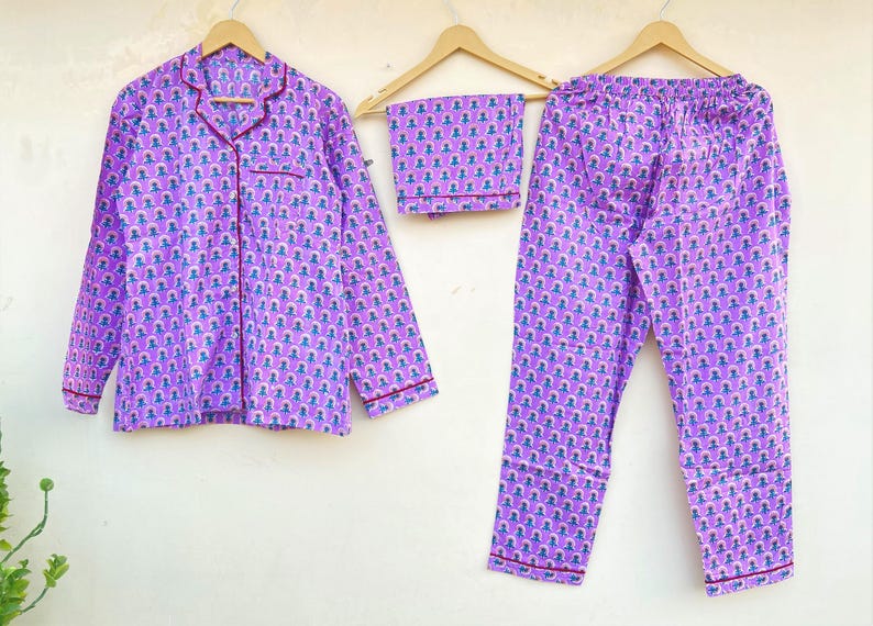 May include: A three-piece pajama set in a lavender hue, featuring a button-down shirt, shorts, and pants. The set is adorned with a repeating pattern of small figures in blue and pink. The edges are trimmed with a dark red piping.