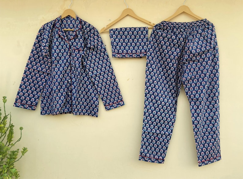 May include: A blue pajama set with a repeating floral pattern in white and pink. The set includes a button-down shirt, pants, and a folded sleep mask. The edges of the shirt and pants are trimmed with red piping.