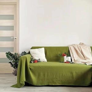 May include: A green sofa with a matching throw, a white cushion, and a beige blanket. Decorative plants and flowers are arranged on the sofa. A light wood door is visible in the background. The room has a neutral colour scheme.