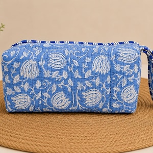 May include: A rectangular quilted zippered pouch featuring a blue and white floral block print pattern. The bag includes a matching blue and white striped fabric wrist strap attached to one side, with blue and white striped piping along the top edge.