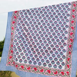 May include: A rectangular textile featuring a central white panel with a repeating blue and pink floral pattern. The center is framed by a pink border with red flowers and green vines, finished with an outer edge of blue and white pinstripes.