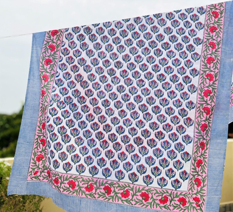 May include: A rectangular textile featuring a central white panel with a repeating blue and pink floral pattern. The center is framed by a pink border with red flowers and green vines, finished with an outer edge of blue and white pinstripes.
