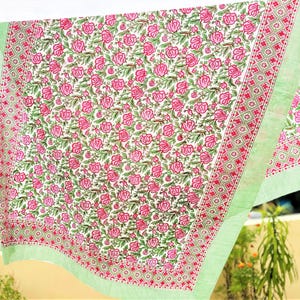 May include: A rectangular textile featuring a central pattern of pink roses with green leaves on a white background. The fabric is bordered by a red and white geometric diamond motif and finished with a light green and white striped edge trim.