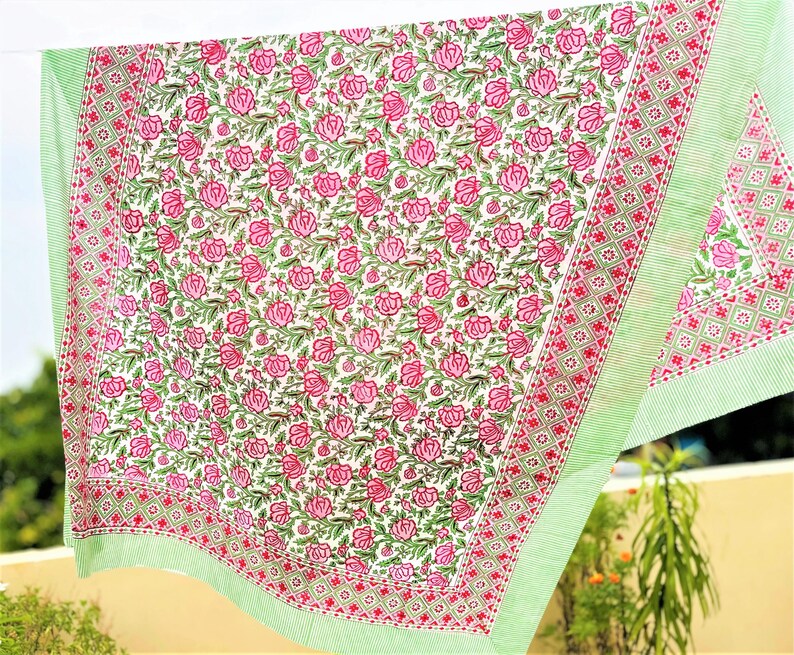 May include: A rectangular textile featuring a central pattern of pink roses with green leaves on a white background. The fabric is bordered by a red and white geometric diamond motif and finished with a light green and white striped edge trim.