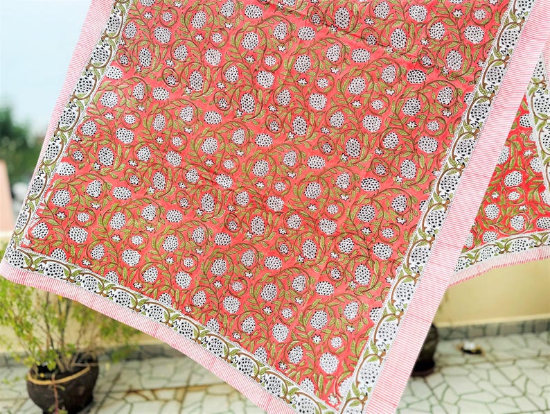 May include: A square textile featuring a vibrant coral-pink background with a repeating pattern of white pomegranate-like fruits, green leafy vines, and small white flowers. The fabric is framed by a border of thin white and coral-pink stripes.