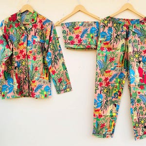May include: A three-piece pajama set with a floral and figure print. The set includes a button-down shirt, shorts, and trousers. The pajamas are pink with a colourful design featuring flowers, figures, and foliage.