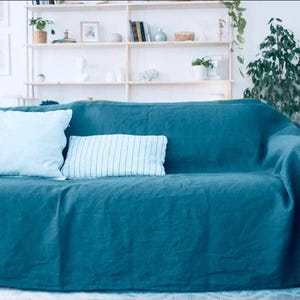 Puede incluir: Un sofá de color verde azulado está cubierto con una manta a juego. Dos almohadas, una lisa y otra a rayas, descansan sobre el sofá. El fondo presenta una pared blanca con estantes que contienen objetos decorativos y plantas, creando un espacio habitable acogedor.