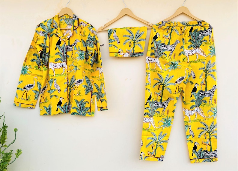 May include: Yellow pajama set with a jungle animal print, including zebras, leopards, and toucans. The set includes a button-down shirt, shorts, and pants, all hanging on wooden hangers against a white wall.