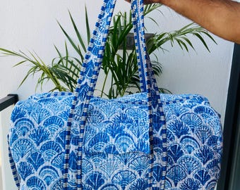 May include: A quilted duffel bag featuring a blue and white fan-shaped shell pattern. The bag includes two long fabric handles with a striped blue and white border, a rectangular front pocket, and a matching adjustable shoulder strap.