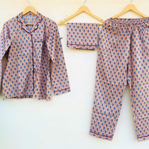 May include: A peach-colored pajama set with a blue and orange floral pattern. The set includes a long-sleeved button-down shirt, a pair of pants, and a matching folded piece. All pieces have red trim and are hanging on wooden hangers.