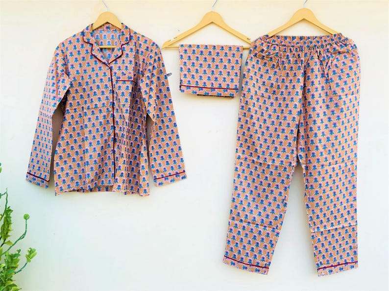 May include: A peach-colored pajama set with a blue and orange floral pattern. The set includes a long-sleeved button-down shirt, a pair of pants, and a matching folded piece. All pieces have red trim and are hanging on wooden hangers.