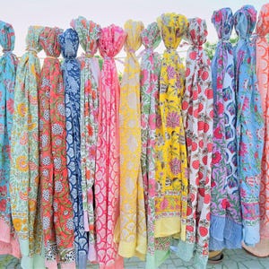 May include: A collection of rectangular fabric scarves hanging vertically, each featuring distinct floral and geometric block-print patterns in vibrant colors including yellow, pink, blue, orange, green, and red, with striped borders at the bottom edges.