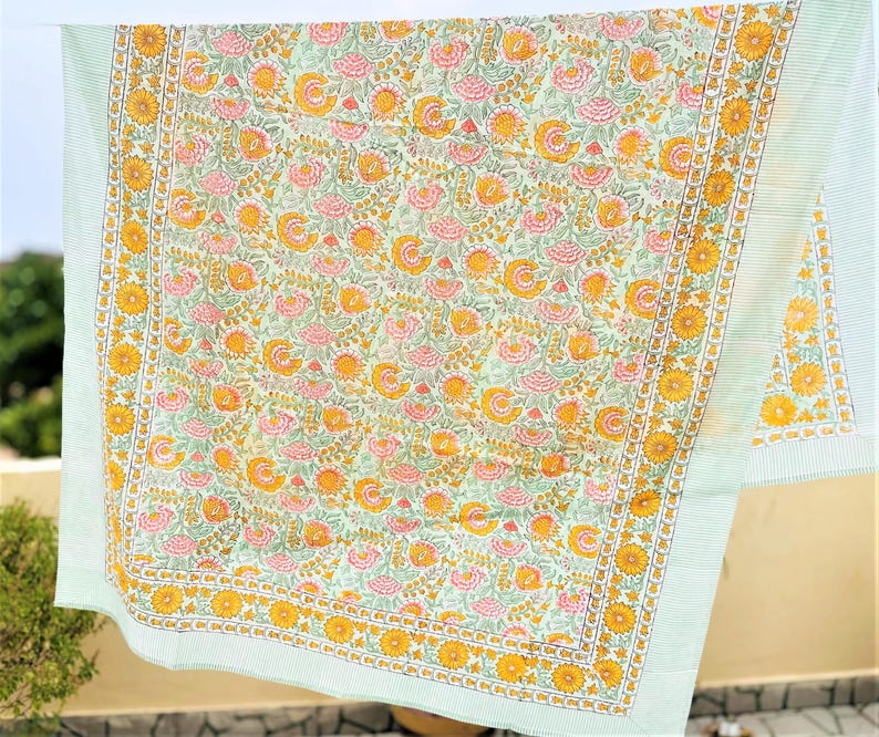 May include: A rectangular textile featuring a light green background with a repeating floral pattern of pink and yellow flowers and green leaves. The fabric is bordered by a wide band of yellow sunflowers and thin light green stripes.