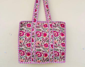 May include: A floral tote bag with a white background and a pattern of pink and orange flowers. The bag has pink trim and handles with a striped design. The bag is made of a quilted fabric.