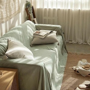 Pode incluir: Um sofá verde claro coberto com um lençol a condizer, complementado por duas almofadas bege com borlas. Um livro aberto repousa sobre uma almofada. A cena é definida numa sala bem iluminada com cortinas de tons neutros e um tapete tecido.