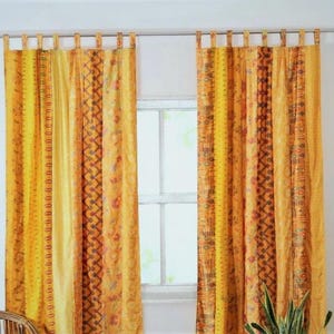 Peut inclure: Deux rideaux à motifs jaunes et oranges suspendus devant une fenêtre. Les rideaux présentent des rayures verticales de motifs et de couleurs variés, notamment des motifs floraux et géométriques. Les rideaux sont fixés à la tringle par des boucles en tissu.