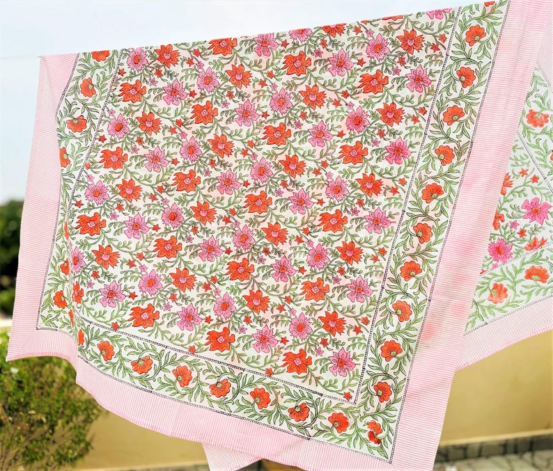 May include: A rectangular textile featuring a repeating floral pattern with orange and pink flowers on a white background. The fabric is bordered by a thin, dark green vine motif and finished with a solid light pink edge on all sides.