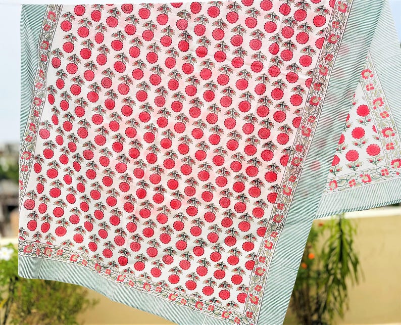 May include: A white fabric textile featuring a repeating pattern of small pink circular floral motifs with green stems. The fabric is bordered by a thin, light green and white striped edge, framed by a decorative floral vine border pattern.