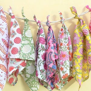 May include: A collection of colorful, patterned fabric napkins hanging from a white string. The napkins feature floral and paisley designs in shades of red, pink, green, and yellow. Each napkin is knotted at the top.