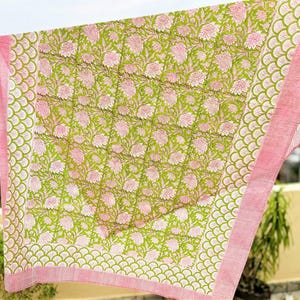 May include: A quilted textile featuring a central panel with a repeating pink floral pattern on a lime green background. The border displays a scalloped green motif followed by a thin pink and white striped edge, finished with visible stitching.