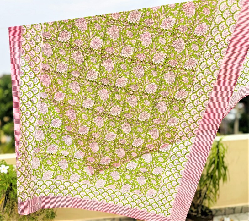 May include: A quilted textile featuring a central panel with a repeating pink floral pattern on a lime green background. The border displays a scalloped green motif followed by a thin pink and white striped edge, finished with visible stitching.