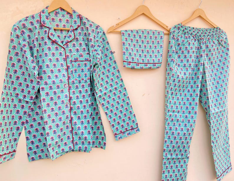 May include: A three-piece pajama set in a floral pattern. The set includes a long-sleeved button-down shirt, pants, and a matching eye mask. The pajamas are light blue with a repeating pattern of pink and blue flowers, and red trim.