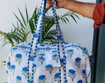 May include: A white and blue floral print duffel bag with a matching shoulder strap. The bag features a quilted texture and blue floral pattern. The handles and strap are trimmed with blue and white stripes.
