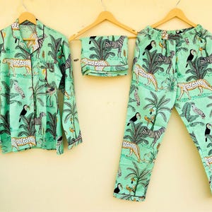 May include: A mint green pajama set featuring a jungle-themed print with leopards, zebras, toucans, and palm trees. The set includes a button-down shirt, pants, and a folded sleep mask, all hanging on wooden hangers.