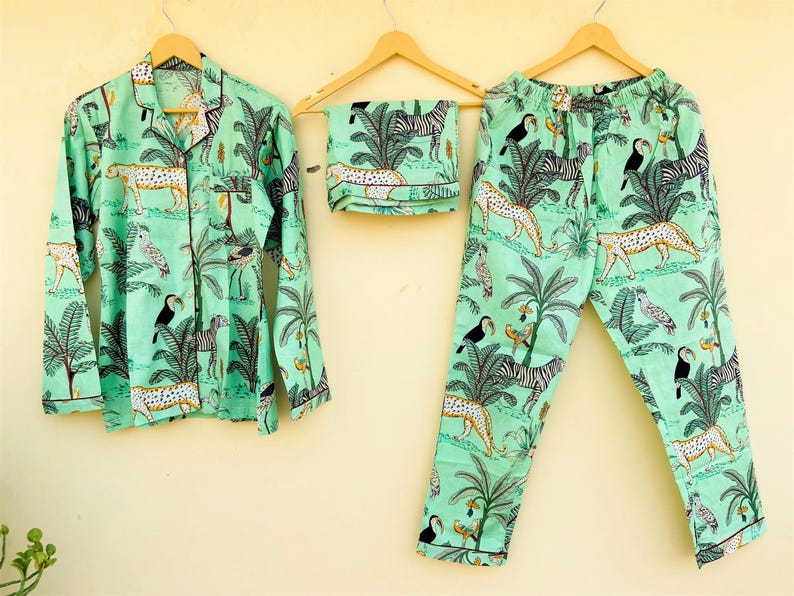 May include: A mint green pajama set featuring a jungle-themed print with leopards, zebras, toucans, and palm trees. The set includes a button-down shirt, pants, and a folded sleep mask, all hanging on wooden hangers.