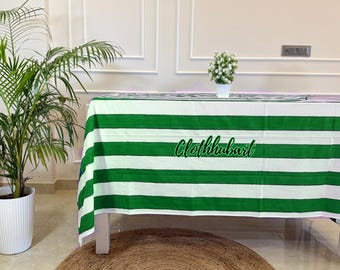 May include: A white tablecloth with bold green horizontal stripes covers a table. The text "Clothhubart" is in a cursive font across the tablecloth. A potted plant and a small floral arrangement are also visible.