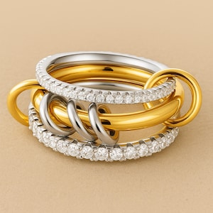Może przedstawiać: Zestaw zazębiających się pierścieni w kolorze złotym, srebrnym i kamieniach przypominających diamenty. Pierścienie są połączone, tworząc unikalny i elegancki design. Biżuteria jest prezentowana na neutralnym tle.