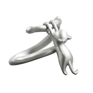 Pode incluir: Anel prateado com um gato estilizado agarrado a uma barra curva. O gato é retratado numa pose brincalhona, com as patas enroladas na barra. O anel tem um acabamento liso e polido e um design minimalista.