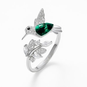 Könnte beinhalten: Silberfarbener Ring mit einem Kolibri-Design. Der Kolibri hat einen grünen Edelsteinkörper und ist mit klaren Kristallen verziert. Ein Zweig mit Kristallakzenten bildet einen Teil des Ringdesigns und schafft eine von der Natur inspirierte Ästhetik.