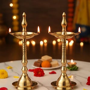 Może przedstawiać: Dwie ozdobne, złote lampy naftowe z zapalonymi płomieniami. Lampy stoją na białej marmurowej powierzchni, otoczone kwiatami i talerzem słodyczy. Tło przedstawia miękkie światła bokeh.