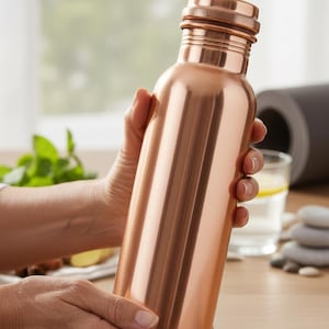 May include: A copper-coloured water bottle with a screw-on cap. The bottle is cylindrical and has a polished finish. The bottle is being held by a person. The background includes a window, a yoga mat, and a glass of water.
