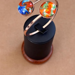 May include: A copper-colored metal bracelet with two circular, colorful, abstract-design cabochons. The cabochons feature a mix of blue, red, orange, and green hues. The bracelet is displayed on a black stand with a wooden base.