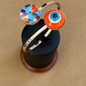 May include: A gold-toned cuff bracelet with two circular charms. One charm features a blue, orange, and white abstract design. The other charm has a red background with a blue and white eye design. The bracelet is displayed on a black stand.