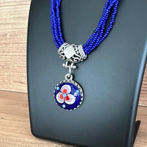 Könnte beinhalten: Eine blaue Perlenkette mit einem runden Anhänger. Der Anhänger hat einen dunkelblauen Hintergrund mit einem Blumendesign in Rot, Weiß und Hellblau. Die Halskette wird auf einem schwarzen Ständer präsentiert.
