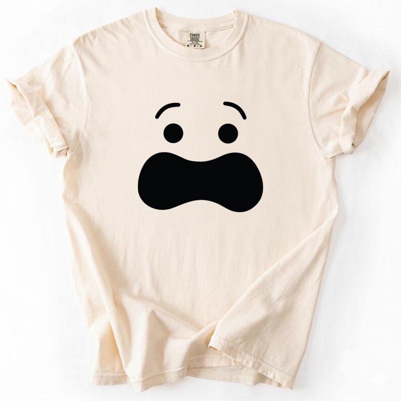 May include: Cream-colored t-shirt featuring a large, black cartoon face. The face has two black circles for eyes and a large, curved black shape for the mouth. The shirt has rolled-up sleeves and a crew neck.