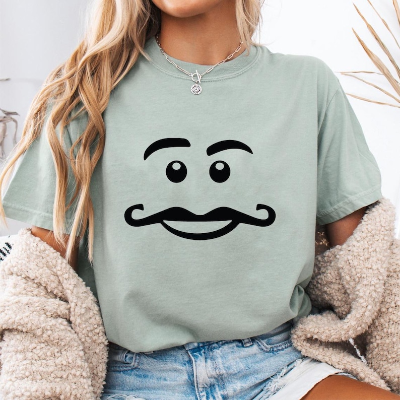 May include: A light green t-shirt featuring a black cartoon face with eyebrows, eyes, and a mustache. The shirt is made of a soft material and is suitable for casual wear.
