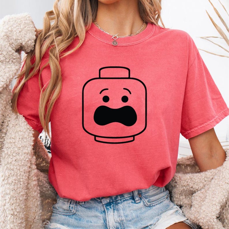 May include: A coral-colored t-shirt featuring a black graphic of a Lego-style face with a surprised expression. The design includes two small eyes and a large, curved mouth. The shirt has short sleeves and a crew neck.