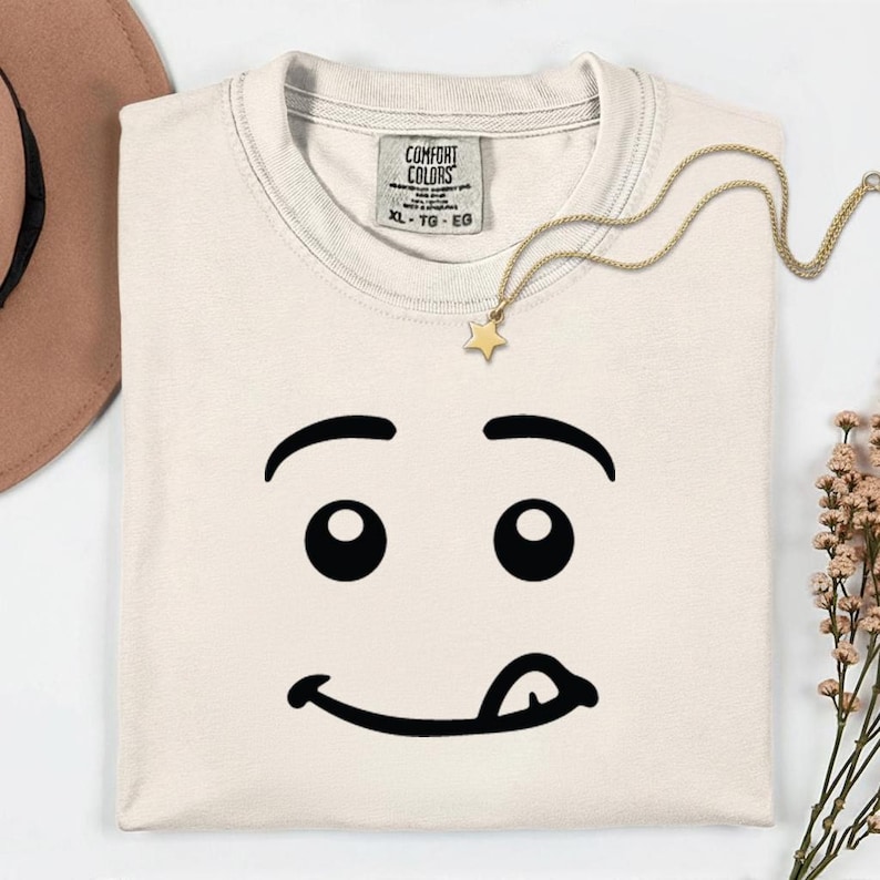 May include: Cream-colored t-shirt with a playful face graphic. The design features black eyes, eyebrows, and a tongue sticking out. A gold star pendant necklace rests on the shirt. The tag reads "COMFORT COLORS" and "XL - TG - EG".