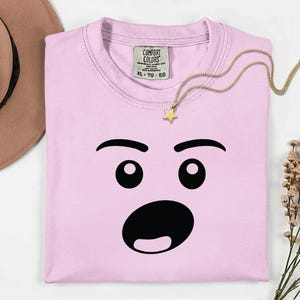 May include: A light pink t-shirt with a surprised face graphic. The graphic features black eyebrows, eyes, and a wide open mouth. A gold necklace with a star pendant rests on the shirt. The shirt has a Comfort Colors tag.