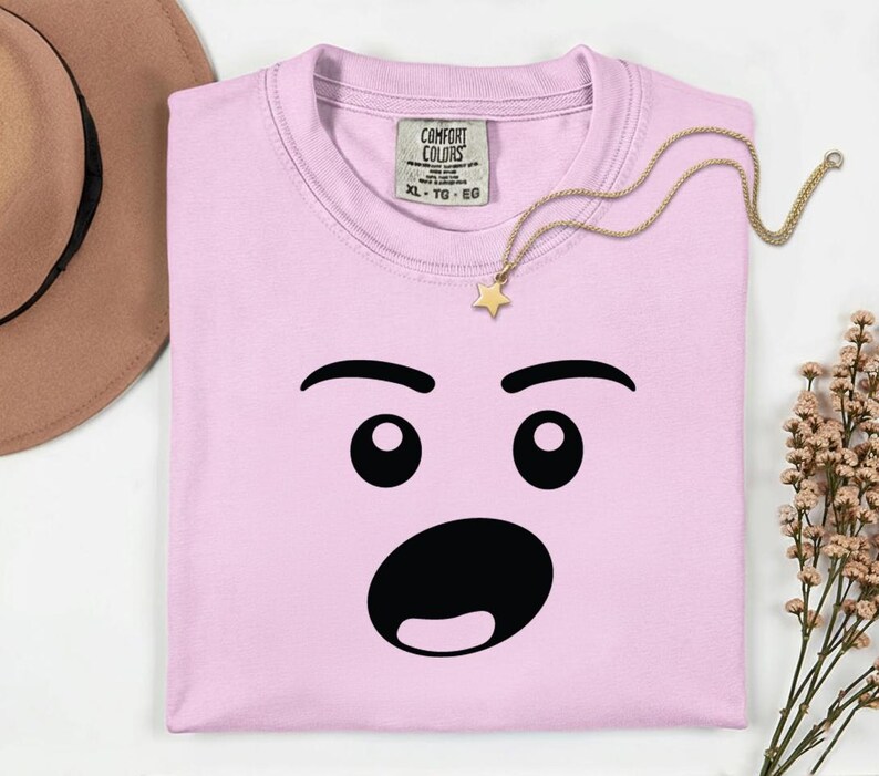 May include: A light pink t-shirt with a surprised face graphic. The graphic features black eyebrows, eyes, and a wide open mouth. A gold necklace with a star pendant rests on the shirt. The shirt has a Comfort Colors tag.